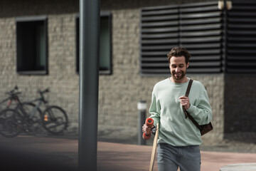 smiling bearded man in sweatshirt holding longboard and leather strap of backpack outside.
