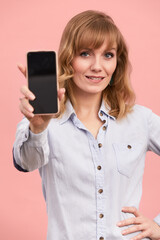 Fototapeta premium Portrait of a girl with a phone in her hands, a beautiful blonde on a pink background, holds a phone in her hands and shows at it, copyspace, pink background