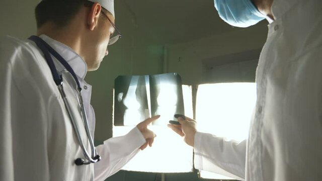 Two caucasian doctors view mri picture and discussing about it. Male medics consult with each other while looking at x ray image. Medical workers in hospital examine x-ray prints. Rear back view