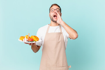 young handsome blonde man feeling happy,giving a big shout out with hands next to mouth. cooking waffles concept