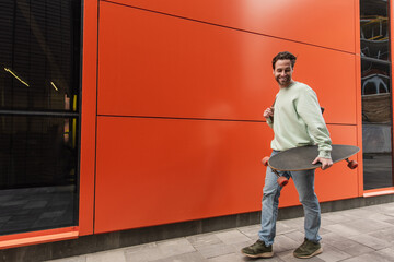 cheerful bearded man in sweatshirt holding longboard while walking near orange wall.