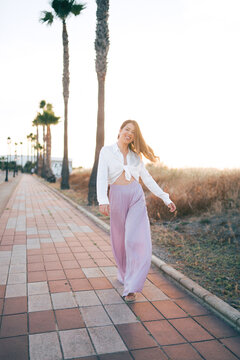 Chica veraniega vestida elegante paseando por zona costera con palmeras 