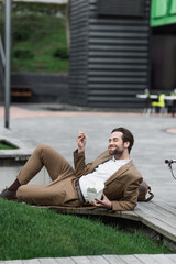 happy businessman holding cardboard box with asian food and chopsticks while lying near grass.