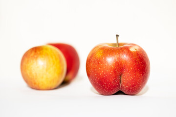 Unusual shape of bright ripe red apple in the shape of an ass in the light of the sun on a white background with two usual apples backdrop. Close-up.  body positive, dissimilarity, uniqueness concepts