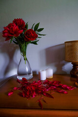 red peonies in glass vase, straw lamp, white candles on wooden commode across white wall. Interior design. Home decor. lights and shadows concept