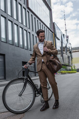 full length of happy businessman in suit holding leather bag and smiling near bicycle outside.