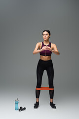 full length view of sportive woman looking away while training with elastics on grey background.