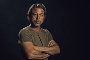 Blond tanned man with a stubble beard in a green t-shirt in front of a dark wall.