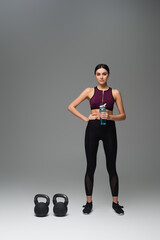 confident woman with sports bottle standing near kettlebells on grey background.