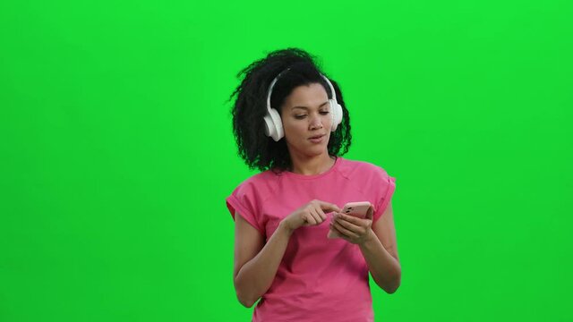 Portrait of young female African American dancing and enjoying music in big white headphones using smartphone. Black woman with curly hair poses on green screen. Close up. Slow motion ready 59.97fps.