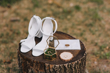 Close-up of the bride's wedding accessories on a textured stump: white leather shoes, gold rings, a...