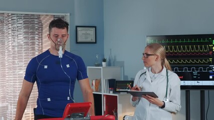 Athlete in oxygen mask doing stress test while cardiologist with tablet monitoring his EKG on display in modern scientific sports laboratory