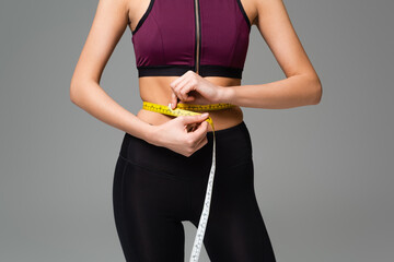 cropped view of woman in black leggings and sports top measuring waist isolated on grey.
