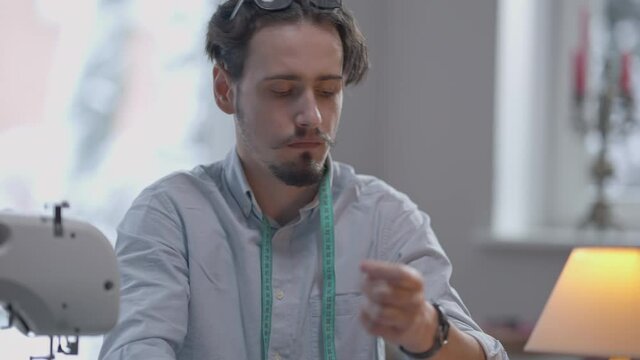 Portrait of young concentrated Caucasian man with beard and mustache threading needle in atelier. Focused confident professional tailor with serious facial expression sewing in studio indoors