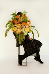 Young lady in black dress sitting on metal stool holding mix of yellow orange color fllowers summer bouquet. 