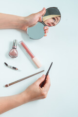 Layout of make up products on white surface and women's hands with cosmetical pencil and hand mirror with reflection of her face. Make up items on the table.