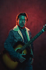 Man with acoustic western guitar in jeans jacket in red and blue colored light.