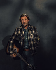 Fototapeta na wymiar Man in checkered jacket with acoustic western guitar in dappled sunlight in front of a grey wall.