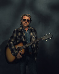 Man in checkered jacket and aviator sunglasses with acoustic western guitar in dappled sunlight in front of a grey wall.