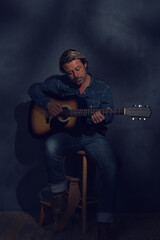 Obraz premium Man in denim shirt and jeans with acoustic western guitar on a wooden stool in dappled sunlight in front of a grey wall.