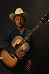 Man in denim shirt and white cowboy hat with acoustic western guitar in front of a grey wall.