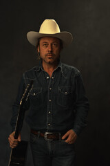 Man in denim shirt and white cowboy hat with acoustic western guitar in front of a grey wall.