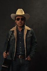 Man in denim jacket, white cowboy hat and aviator sunglasses with acoustic western guitar in front...