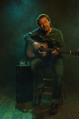 Obraz premium Musician sitting on a wooden stool plays an acoustic guitar with next to him a guitar case with a glass of whiskey and an ashtray on it.