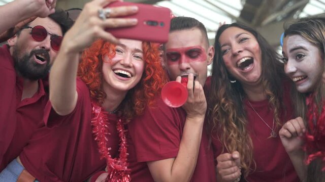 Excited sport fans taking a selfie after the victory of their team in the soccer game - Young group of happy multi ethnic friends having fun at football stadium