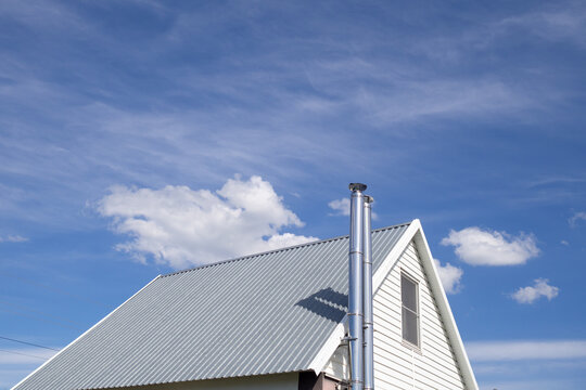 White metal roof of cottage with stainless steel pipe against blue sky. Roof is made of galvanized metal profile.