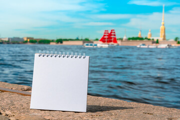An empty white notebook with space for text stands on the Neva River embankment in St. Petersburg in the summer, mock up