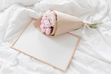 White frame mockup on the bed. Bouquet of pink peonies in craft packaging. Scandivanavian white...