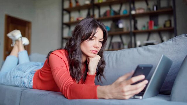 Side view of attractive woman lying on couch at home and working on laptop computer. Female answering call on cell phone, talking, finishing conversation and proceeding work
