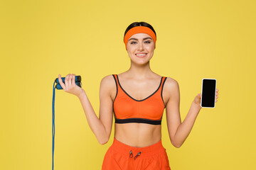 sportswoman with skipping rope and smartphone with blank screen smiling at camera isolated on yellow.