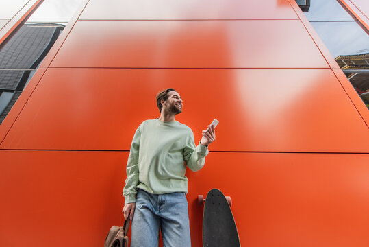 low angle view of cheerful man in sweatshirt holding backpack and smartphone near longboard and orange wall.
