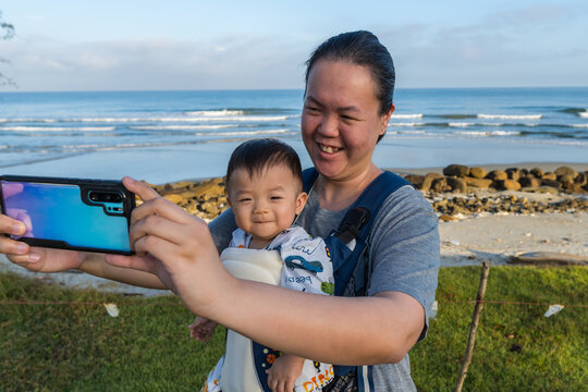 Local lifestyle Asian Chinese mother carry her baby while talking selfie photo with baby on beautiful tropical beach.