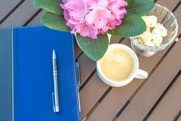 A tablet with a pen and in a vase white chocolate and an alpine rose on a wooden table and a cup of coffee with sweets,