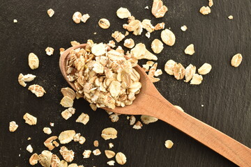 Organic uncooked oat flakes with a wooden spoon on a slate board, close-up, top view.