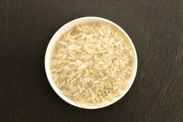 White uncooked rice in a white dish, close-up, on a slate board, top view.