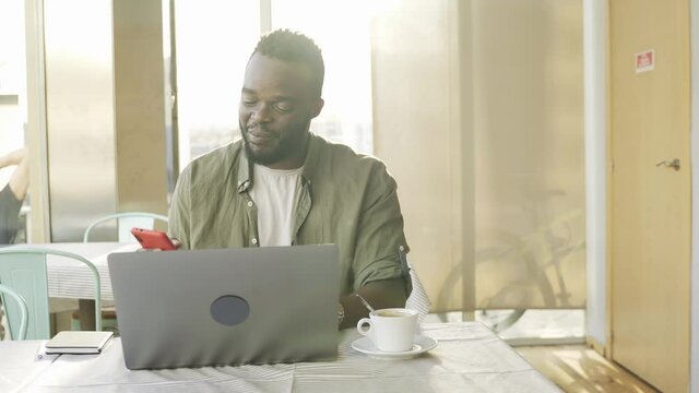 Young african man using mobile phone and computer laptop while working at cafe bar restaurant