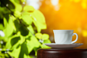 White cup of coffee or tea on a wooden table over blurred tree with sun lighting
