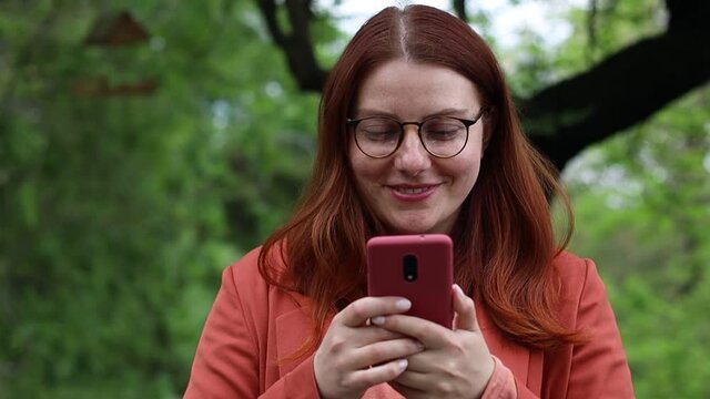 Smiling young caucasian millennial girl in eyeglasses holding smartphone using mobile application technology outdoors. Happy young woman texting checking online social networking applications in city