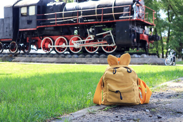 The concept of summer travel by rail. Yellow backpack on the background of an old steam locomotive on a sunny summer day, close-up