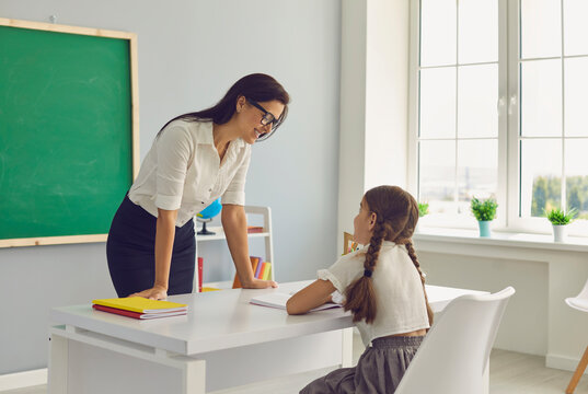 Smiling young female teacher tutor and smart girl elementary school pupil talking in classroom. Student sitting at desk listening to educator. Education and back to school, elective lesson study group