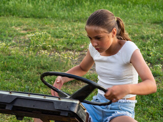 Obraz premium A beautiful teenage girl turns the steering wheel on a makeshift car on a green lawn. A scene from real life