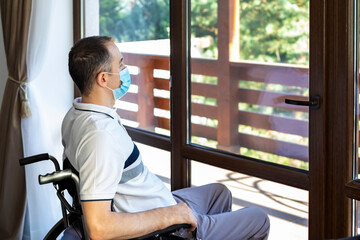 young man wearing face mask sitting in a wheelchair alone looking out the window