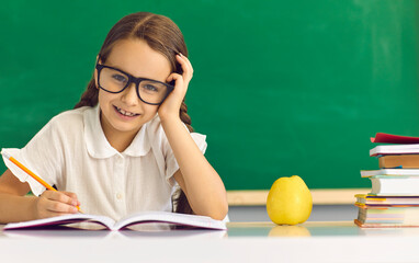 Happy elementary school girl student headshot portrait. Female pupil wearing eyeglasses with pen writing in copy-book sitting at desk and studying against green blackboard. Back to school concept