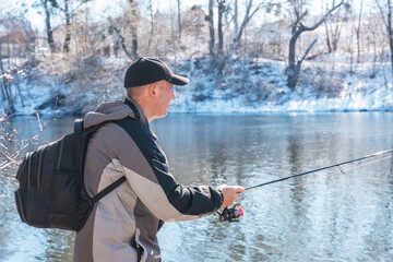 A fisherman with a fishing rod and a backpack catches fish on the bank of a snow-covered river in early spring