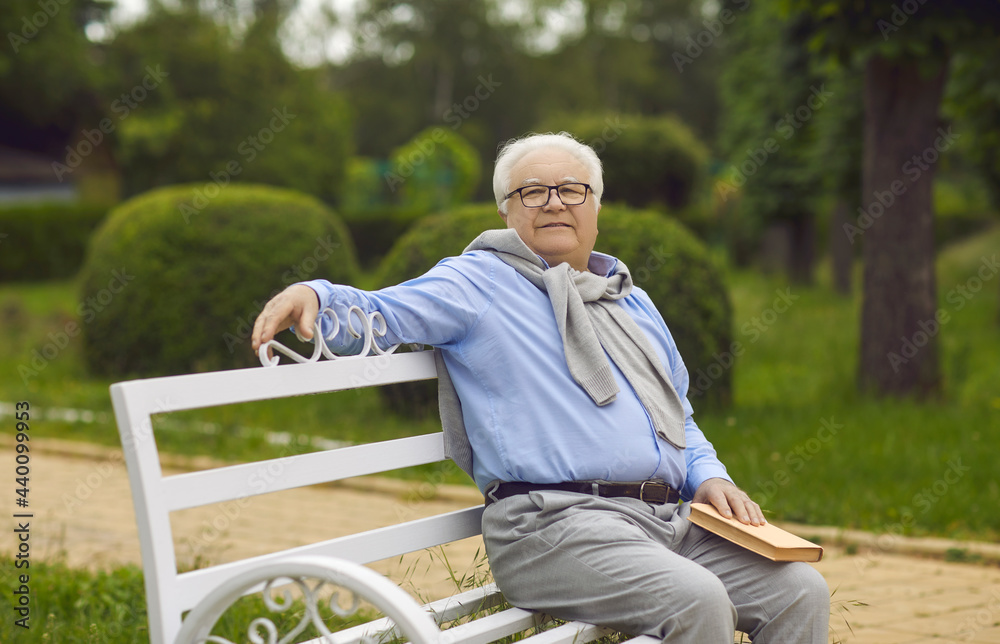 Wall mural senior man sits on a bench and reads a book. an elderly man with white hair is resting on a park ben