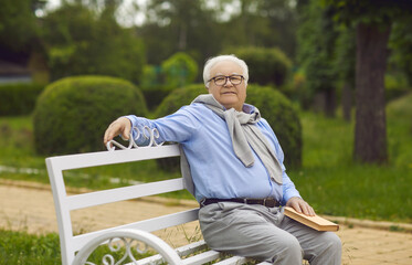 Fototapeta premium Senior man sits on a bench and reads a book. An elderly man with White hair is resting on a park bench. Concept of happy elderly man reading books in old age.